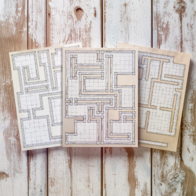 Three hand-drawn dungeon battle maps printed on textured paper, displayed on rustic wooden boards.